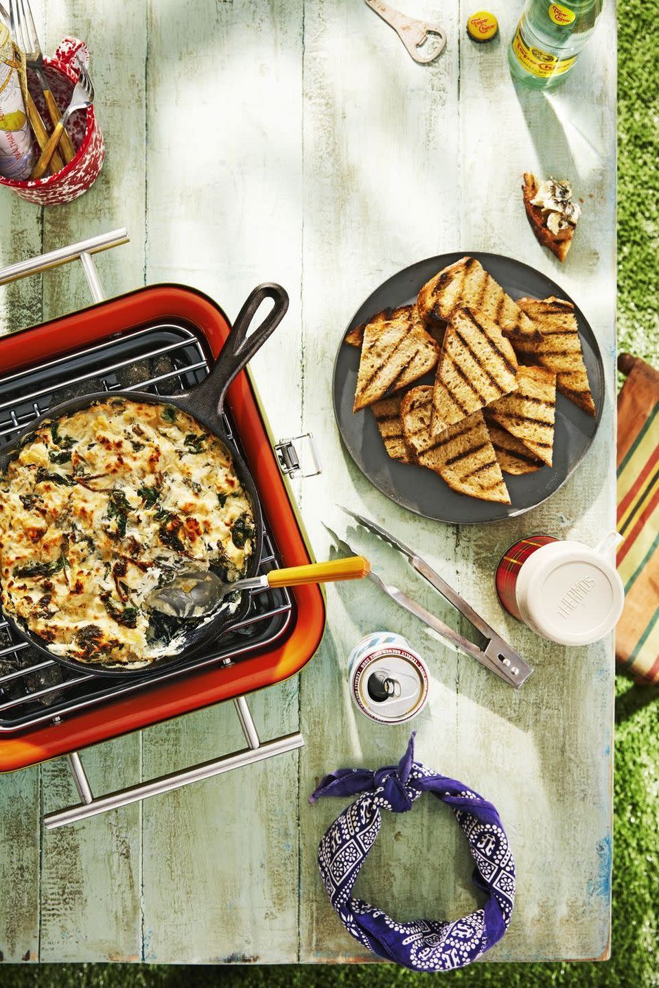 skillet spinach artichoke dip in a cast iron skillet on a camp grill with fire roasted bread on a plate to the side