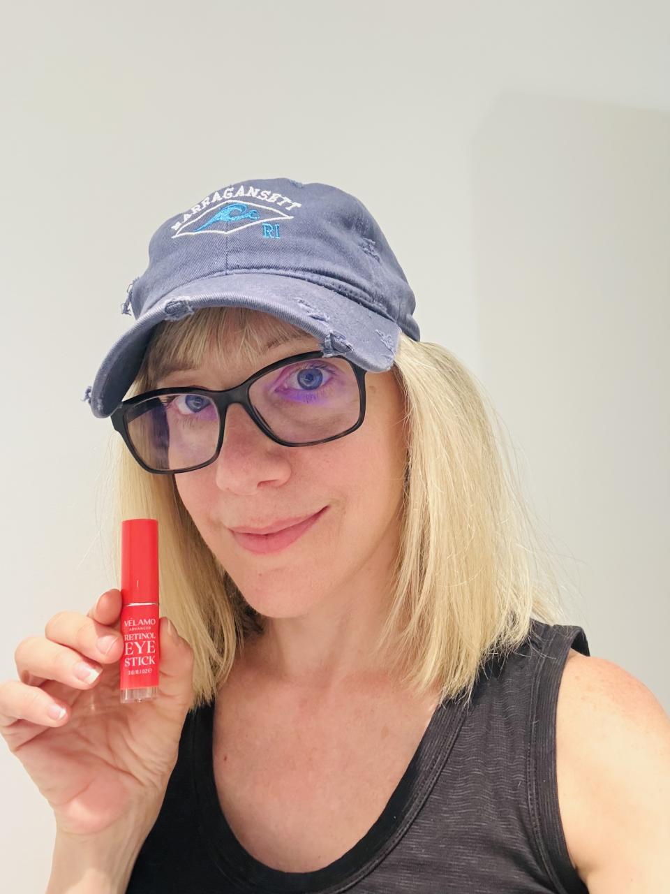 blonde woman in baseball cap and black tank top holding velamo retinol eye stick