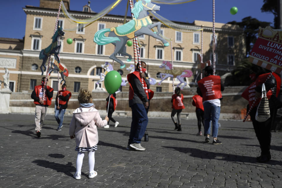 A child holds a balloon as she looks at amusement park workers holding a makeshift carousel as they join with circus workers in a demonstration demanding more support from the Italian government as their activities are shut down since the start of the COVID-19 outbreak, in Rome's Piazza del Popolo, Friday, March 26, 2021,. (AP Photo/Gregorio Borgia)