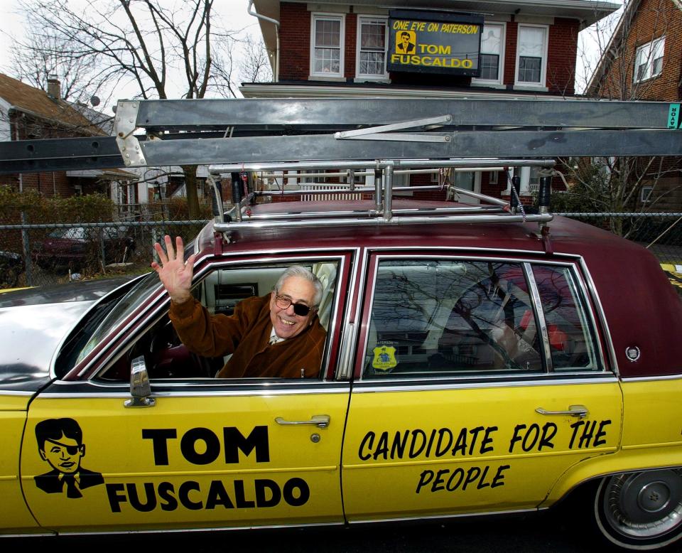 Tom Fuscaldo waves from the front seat of his Cadillac, with his logo painted on the side, and a ""One Eye on Paterson"" sign on the upper part of the house, January 7, 2004.