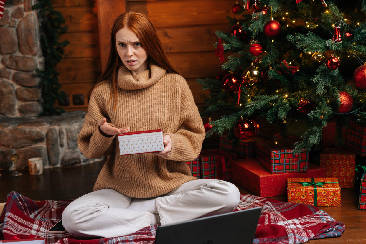 Happy excited young woman waiting for good Christmas present, lady with disappointment opening box with surprise on background of xmas tree at living room. Female looking at camera incomprehensibly.�