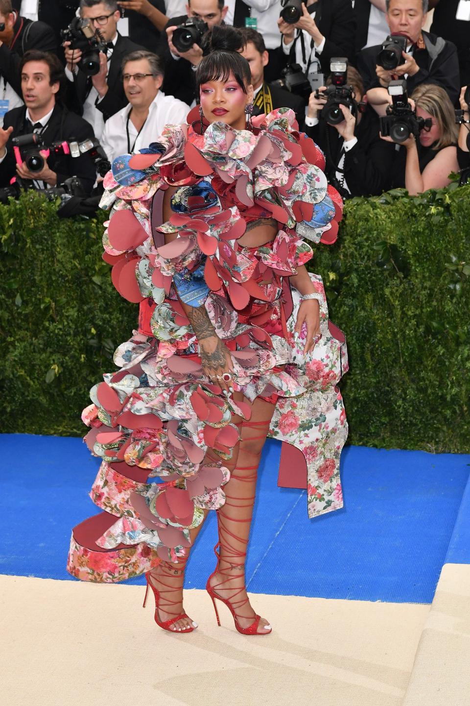 Rihanna poses for photos on a previous Met Gala red carpet in strappy red heels and a sculptural look made from colorful floral fabrics.
