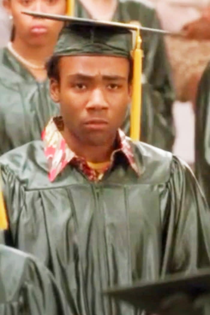 Donald Glover in a green graduation gown and cap stares seriously, surrounded by other graduates, in a scene from 