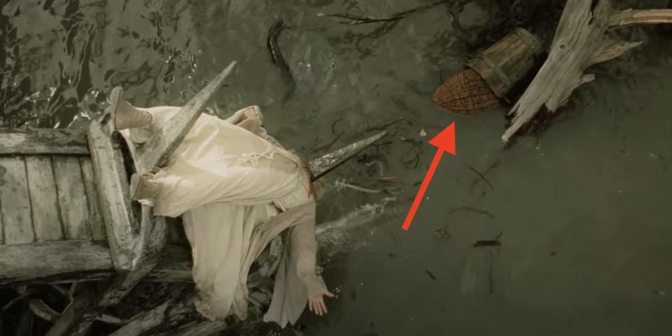 The body of Saruman (a white-robed wizard) impaled on a wheel that is slowly turning into the water. A nearby basket is floating on top of the water.