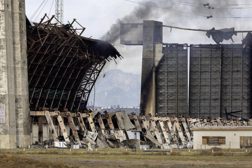 Tustin, CA - November 13: A disaster cleanup crew picks up potentially toxic debris around the still-burning WWII-era blimp hangar at the former air base in Tustin Monday, Nov. 13, 2023. Flare-ups and toxic air from last week's destructive hangar fire in Tustin continue to cause trouble for nearby residents. The City of Tustin took to X, formerly Twitter, to confirm that the western wall of the 17-story building reignited Sunday night. Orange County Fire Authority personnel remained on the scene keeping watch of the blaze on Monday morning, with one firefighter telling KTLA 5's Annie Rose Ramos that all they could do was let it burn out. (Allen J. Schaben / Los Angeles Times)