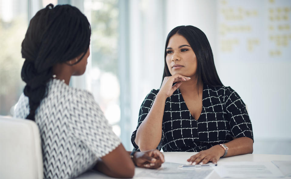 Two women are having a serious conversation at a table in a bright office setting. One woman is facing the camera, and the other is seen from behind