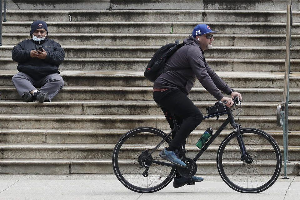 A man checks his phone as a man rides bicycle on the sidewalk in downtown Chicago, Wednesday, April 22, 2020. Illinois Gov. Jay Pritzker said the state could reopen in stages, with each region facing different restrictions amid the COVID-19 pandemic. (AP Photo/Nam Y. Huh)