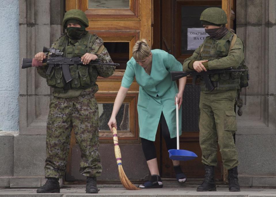 A woman sweeps away the broken glass as two unidentified armed men guard the entrance to the local government building in downtown Simferopol, Ukraine, on Sunday, March 2, 2014. A convoy of hundreds of Russian troops headed toward Simferopol the regional capital of Ukraine's Crimea region on Sunday, a day after Russia's forces took over the strategic Black Sea peninsula without firing a shot. (AP Photo/Ivan Sekretarev)