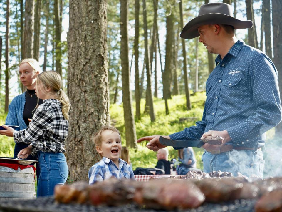 A man in western style clothes and a cowboy hat cooking next to a young child in the woods, and two other people.