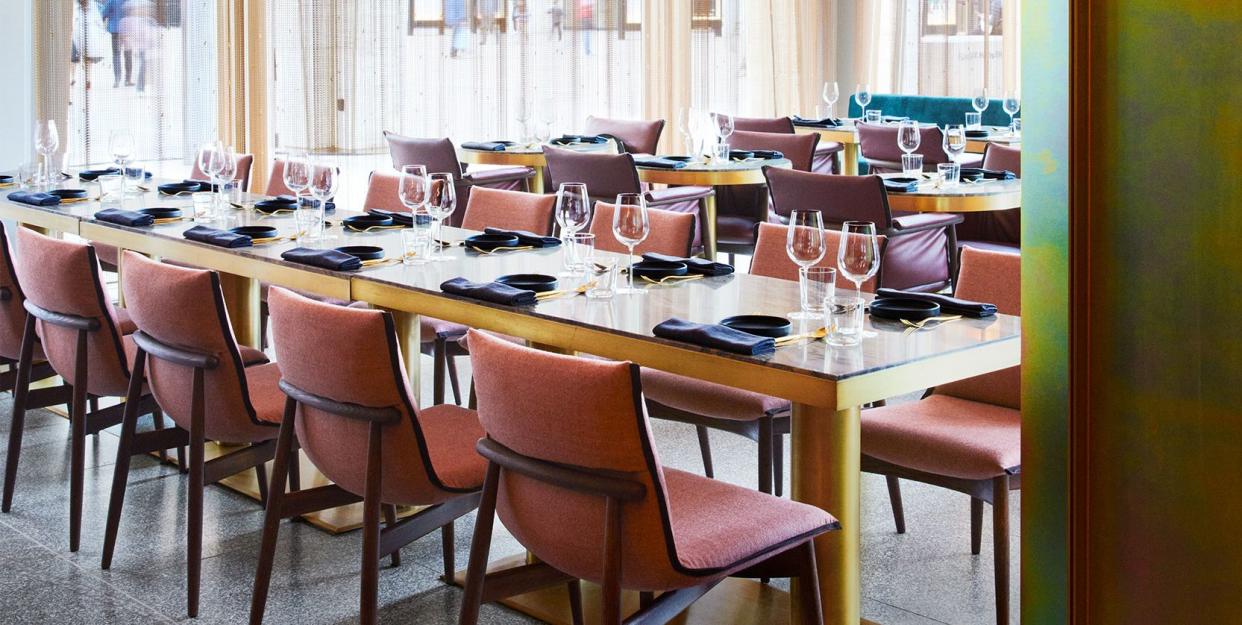 interior of restaurant  with a long table set with armless scoop back chairs and tall windows with sheer curtains and large white floating fixtures hanging from the ceiling