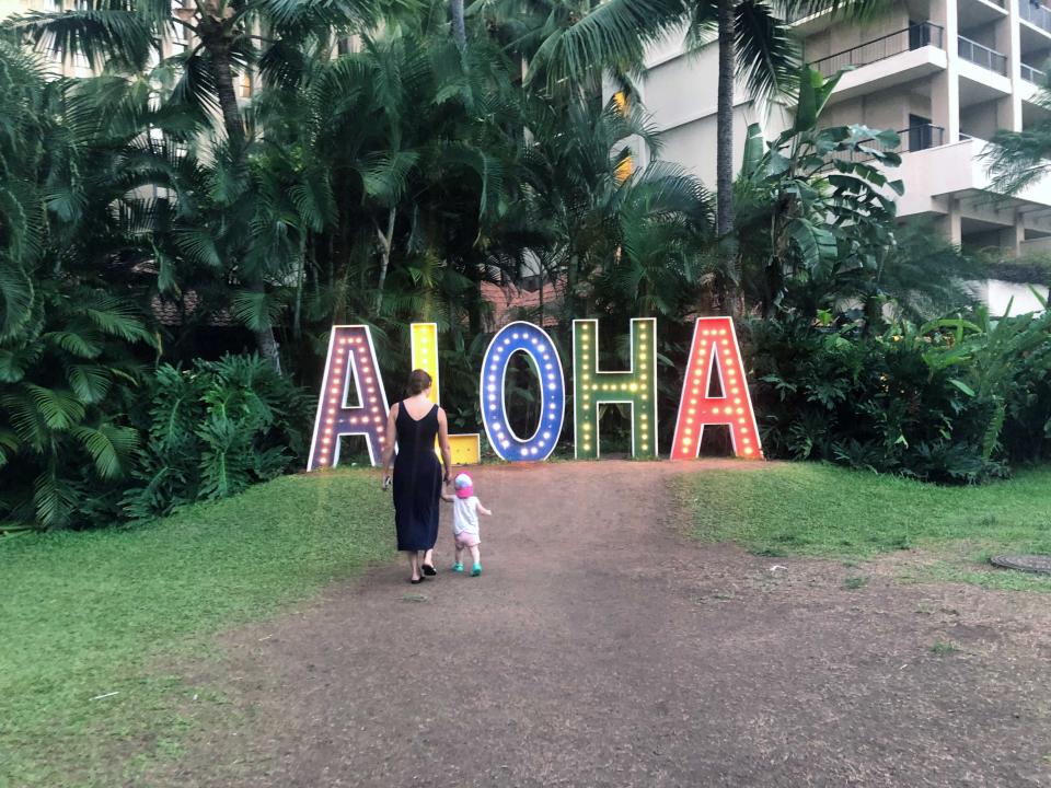 A woman walking holding the hand of her small child to a lit up sign that says Aloha.