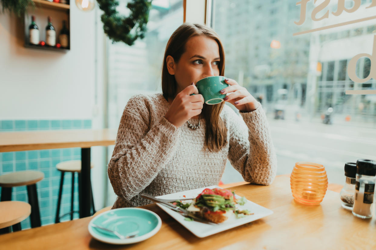 Young woman sitting in the coffee bar, solo dining, drinking coffee from a blue mug and eating healthy vegan avocado toast for breakfast