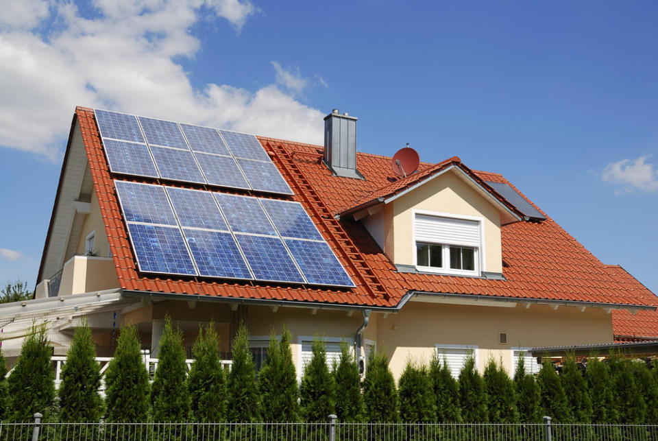 A house with a red roof topped with solar panels against a blue sky with minimal clouds.