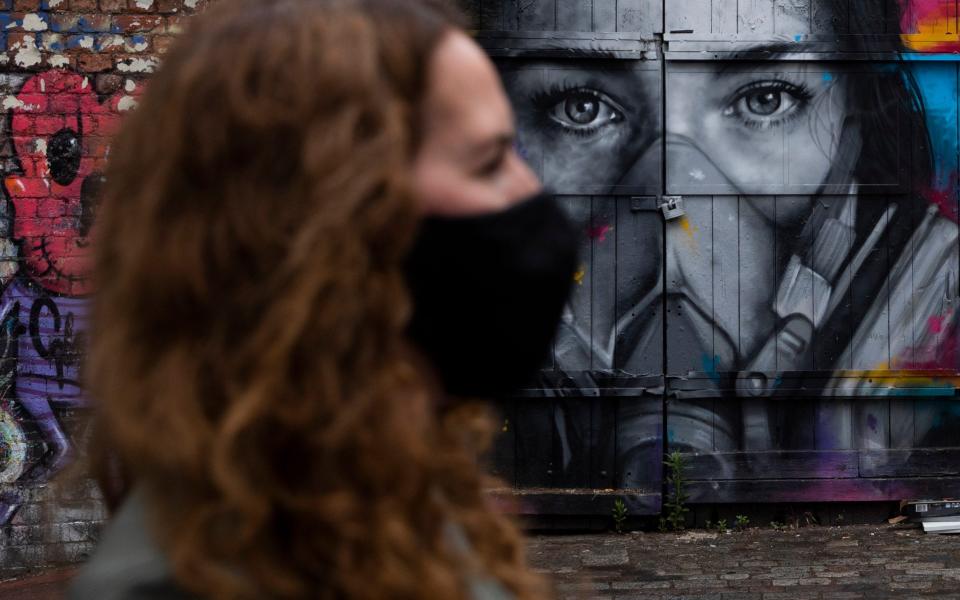 A woman wearing a face mask walks past a mural of a woman wearing a face mask in Shoreditch, east London - Getty Images