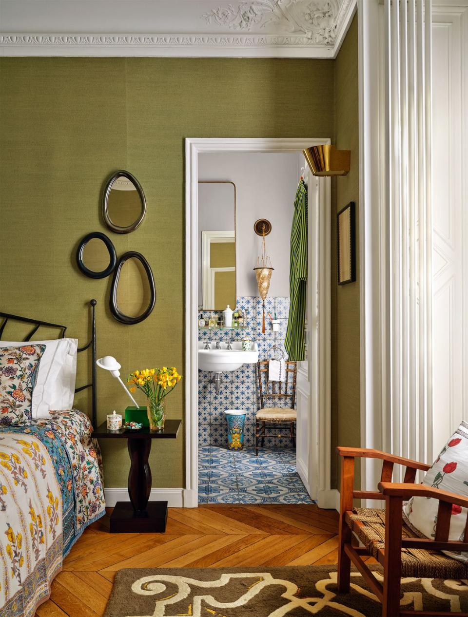 the side of bedroom has an iron bed, a nightstand with three oval mirrors above it, an armchair, a brown patterned rug, a door to the bathroom with tiled floor and walls, a sink, cone sconce and mirror