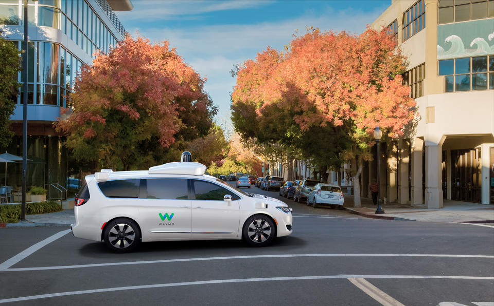 Waymo self-driving minivan crossing an intersection with fall trees in the background.