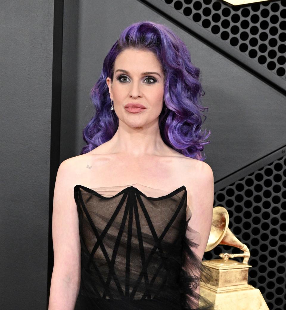 Kelly Osbourne, with long purple hair, in a strapless black gown with sheer overlay at the Grammys; she carries a clutch with a recording sleeve design