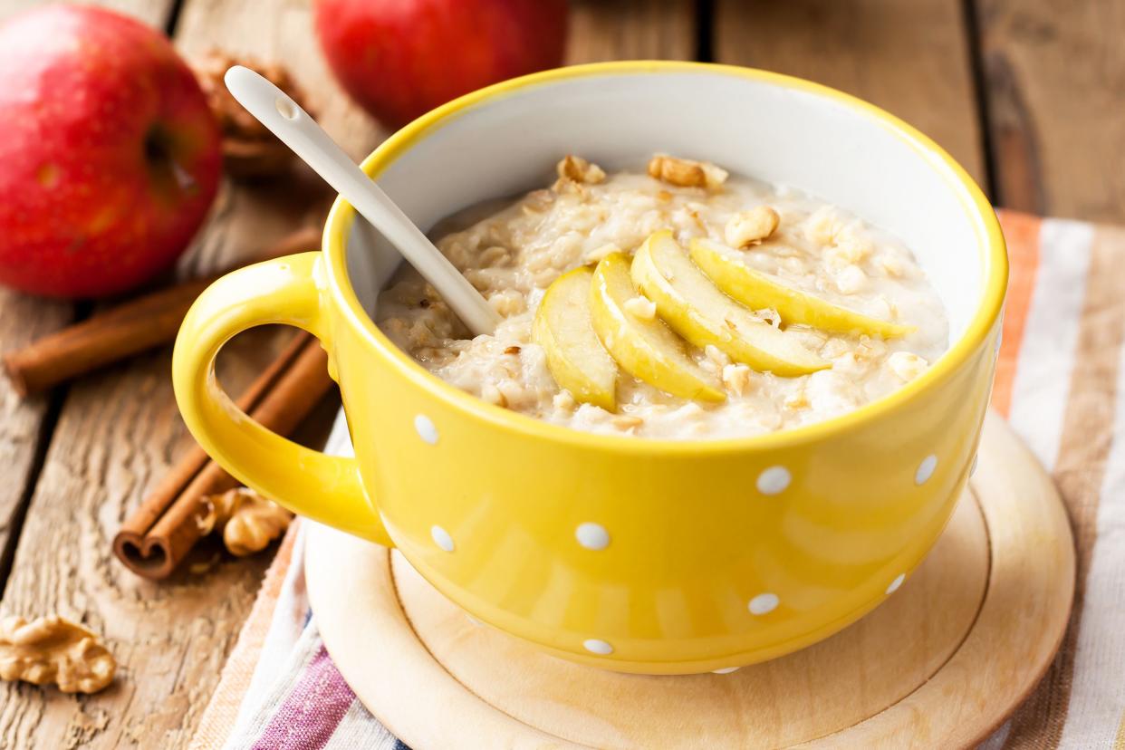 Apple walnut oatmeal with a spoon in a yellow mug with white polka dots on a beige saucer, on a striped napkin, surrounded by ingredients