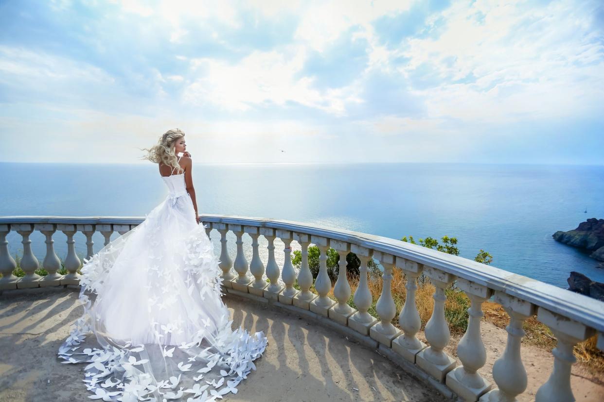 Bride in a luxurious and fancy white wedding dress, standing on a balcony overlooking the ocean on a sunny summer day