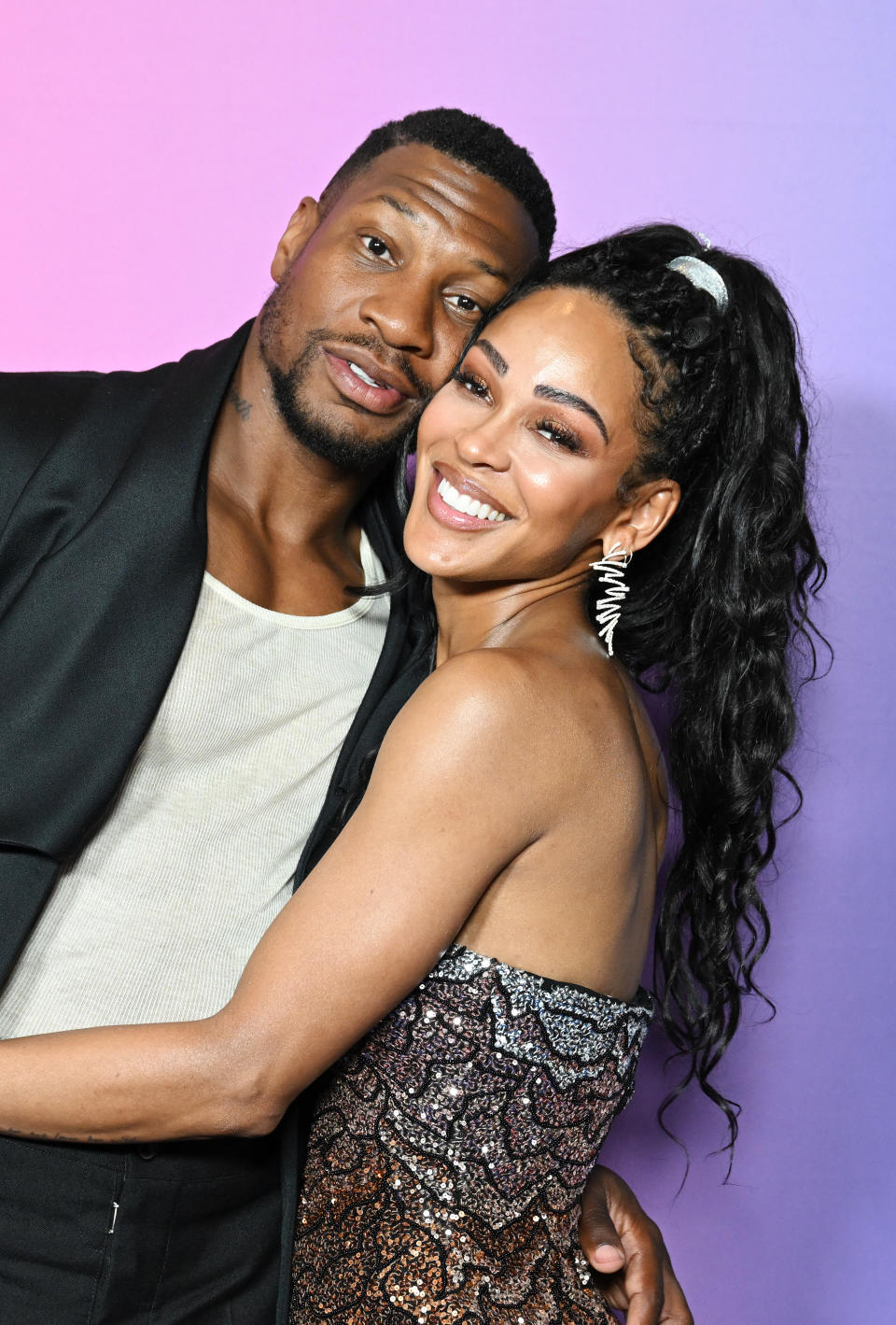 Jonathan Majors and Meagan Good smile and hug closely. Jonathan wears a casual top with a jacket, and Meagan wears a strapless, patterned dress with statement earrings