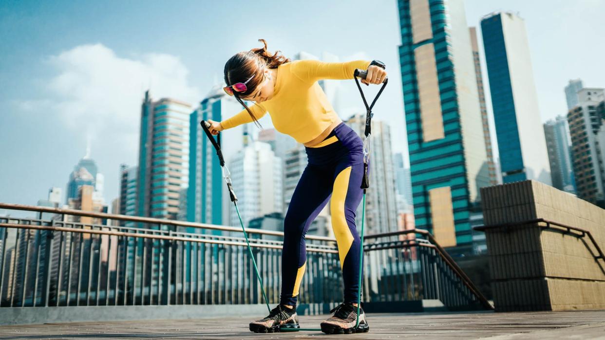 Young Asian sports woman exercising with a resistance band outdoors, with urban city skyline as background.