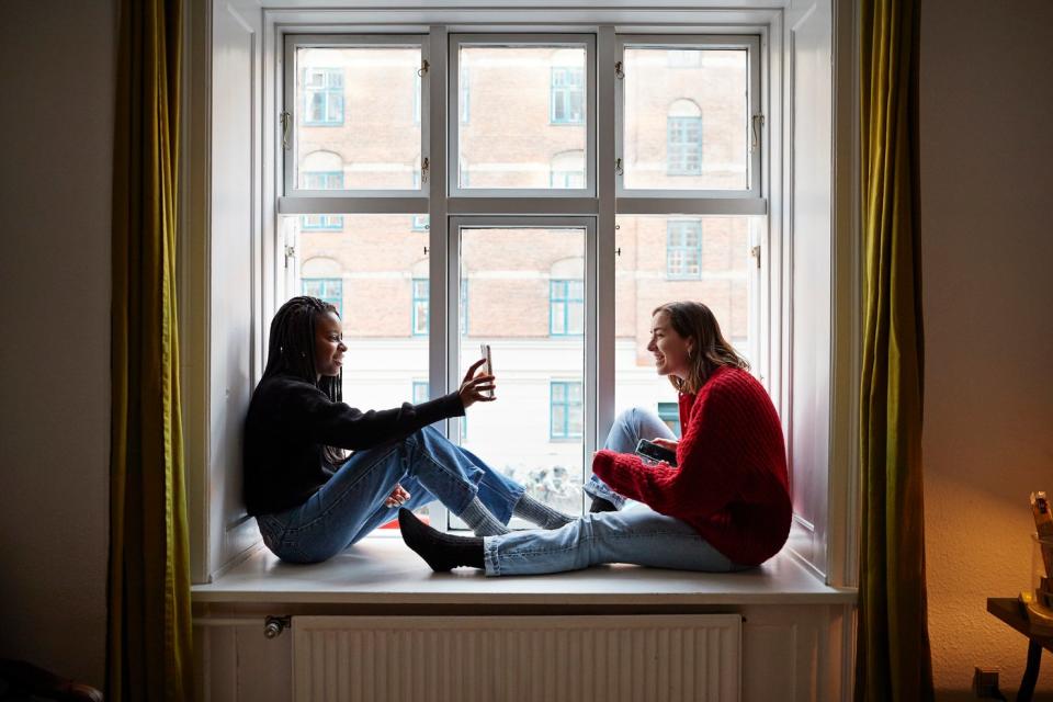 Two friends sharing a smartphone screen by an apartment window.