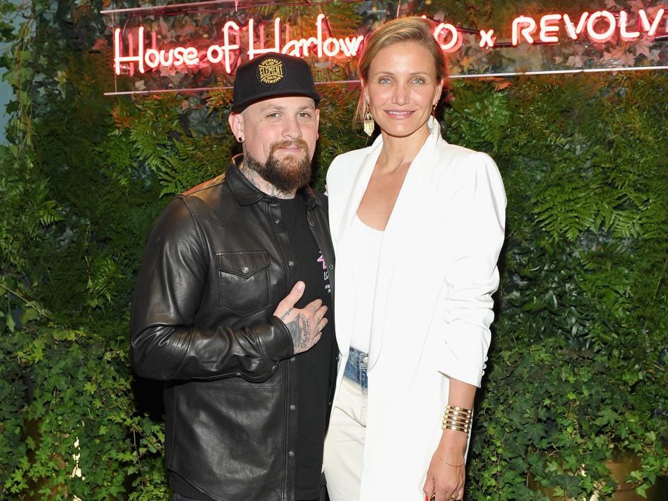 Cameron Diaz in a white blazer and pant ensemble and heels standing taller than Benji Madden in a leather jacket, hat, and Converse sneakers.