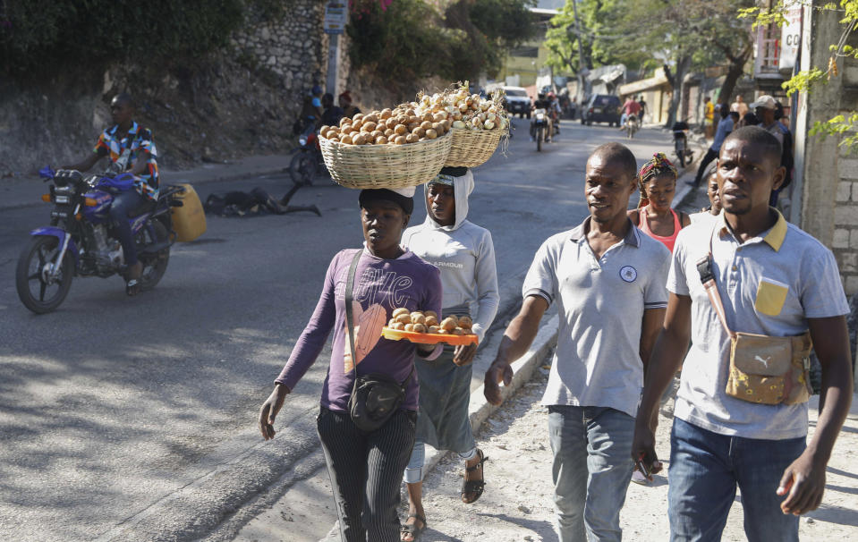 Motociclistas esquivan un cadáver calcinado que yace en el camino mientras varios peatones pasan a su lado en el vecindario de Petion-Ville de Puerto Príncipe, Haití, el miércoles 20 de marzo de 2024. (AP Foto/Odelyn Joseph)