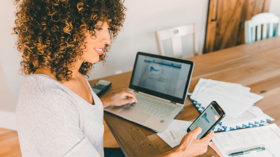 A woman sits at her dining room table with laptop and financial reports doing her monthly budget.