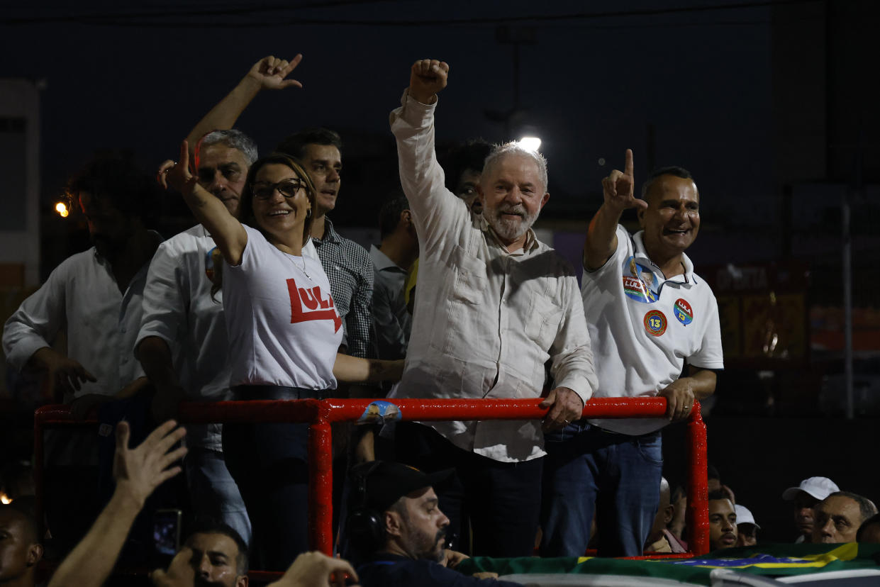 Luiz Inácio Lula da Silva, with a white beard and a shirt with no tie, raises his hand in a fist, surrounded by four associates making the L sign, for Lula, inside a red metal rail.