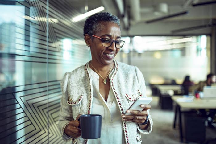 A woman with short cropped hair and eyeglasses standing in an office holding a mug and looking at her phone