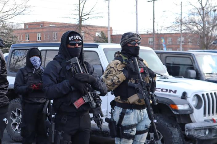 In dark glasses with masks and wool hats, and carrying rifles, members of New Era stand guard on a city street.