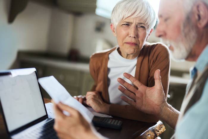 An older couple is sitting at a table, appearing frustrated while discussing paperwork and looking at a laptop screen