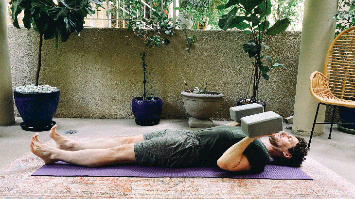 May lying on his back on a yoga mat with his elbows bent and foam blocks balancing on his hands in a variation of Chaturanga