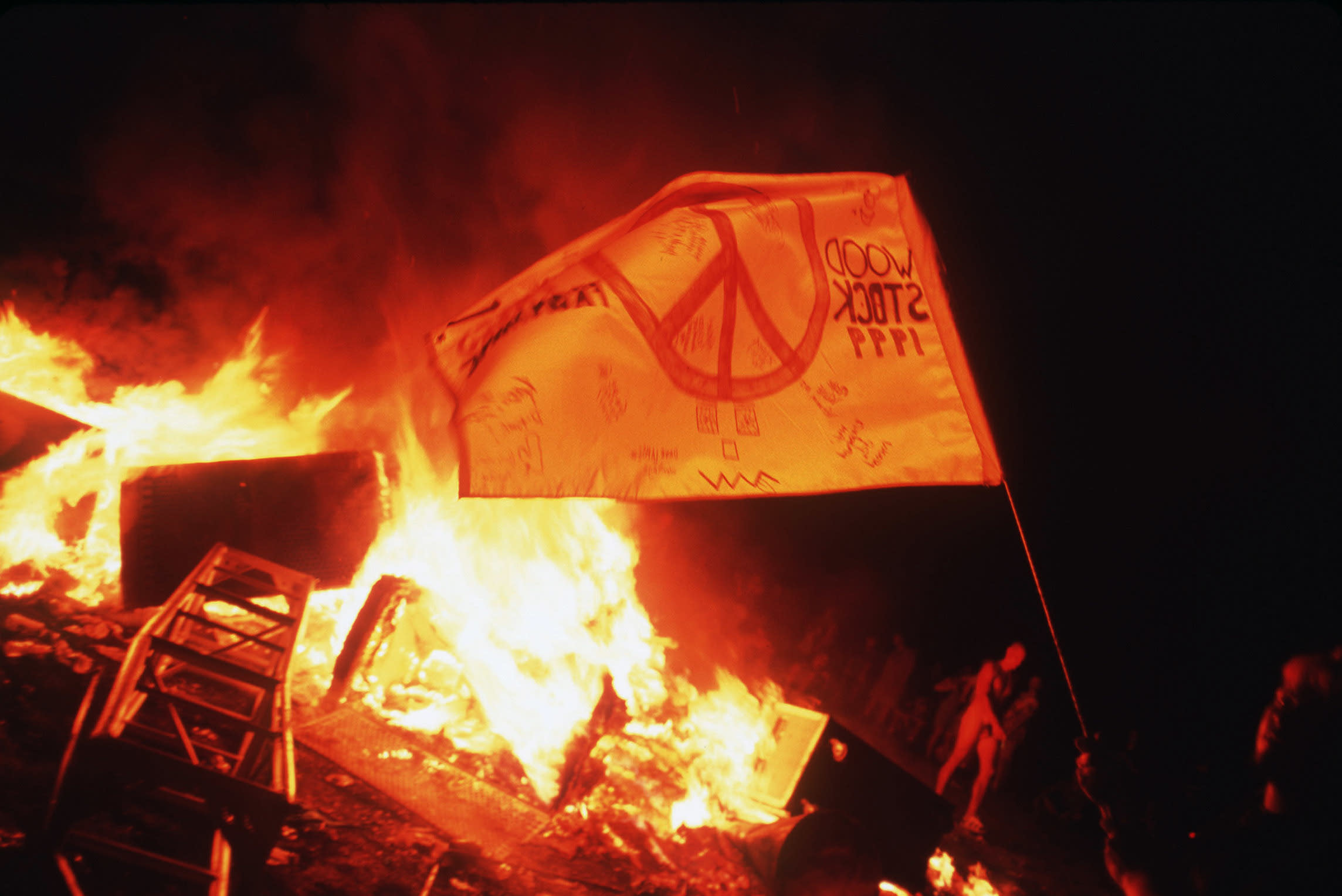 A flag with a peace sign on it before a raging fire.