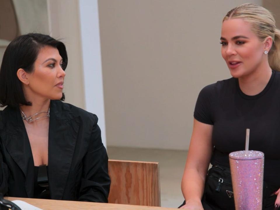 kourtney kardashian sits at a table, looking sideways to her sister khloé, who is sitting at the table and speaking