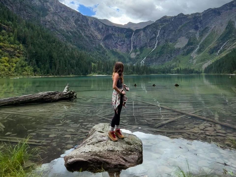 Emily stands on a rock in the middle of a lake with clear water in Glacier National Park. The lake is surrounded by mountains and trees.