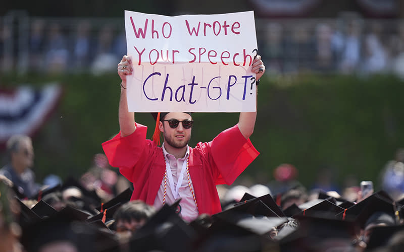 A person in a graduation cap and gown holds a sign that reads "Who wrote your speech? ChatGPT?" during an address by the president and CEO of Warner Bros. Discovery