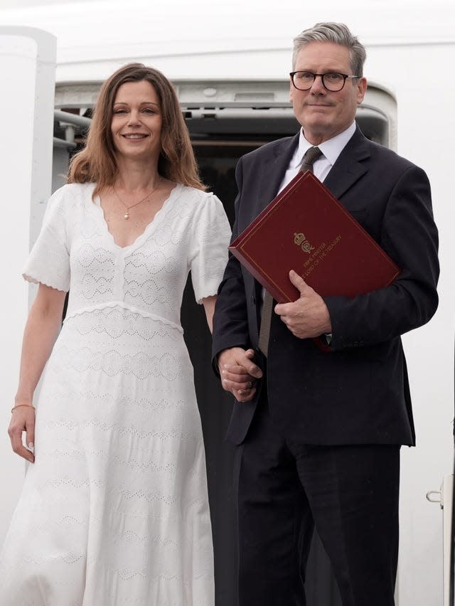 Prime Minister Sir Keir Starmer and his wife Victoria standing on the steps of a plane, with Lady Starmer wearing a white dress and Sir Keir holding a burgundy folder