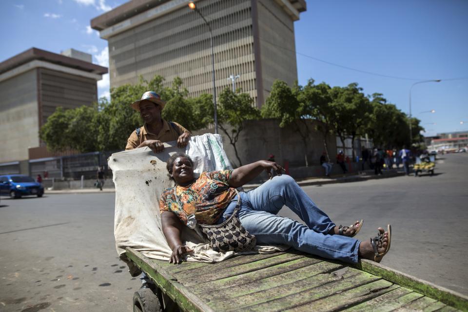 In the Nov. 19, 2019 photo, Neyda Rosa Hernandez poses for a photo with her husband Osmar Fernandez while reclining on the cart she uses to sell bananas, in Maracaibo, Venezuela. Pictured in the background are offices of the state oil company PDVSA. Critics blame two decades of socialist rule for destroying the oil industry, that today produces a fraction of what it did at its height two decades ago. The Venezuelan government blames U.S. sanctions for many of its problems. (AP Photo/Rodrigo Abd)