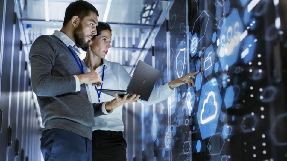 Male IT Specialist Holds Laptop and Discusses Work with Female Server Technician. They're Standing in Data Center, Rack Server Cabinet with Cloud Server Icon and Visualization