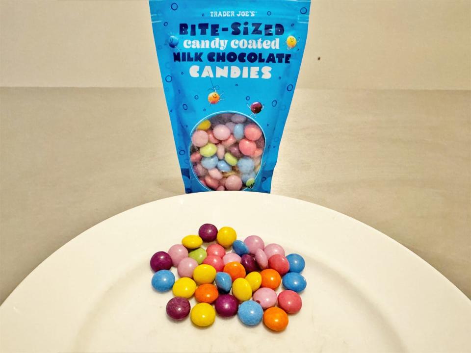 Blue bag with see-through window of Trader Joe's bite-sized candy-coated milk-chocolate candies with multicolored candies on plate in front