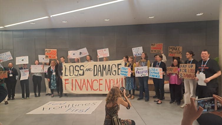 grey room with more than a dozen young people holding climate protest signs, photographer in front of them kneeling down take a pic of them