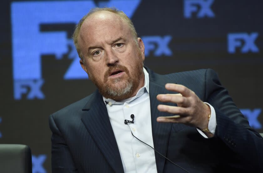 Louis C.K.in a black suit jacket, white suit shirt sitting and holding his left hand out infant of him