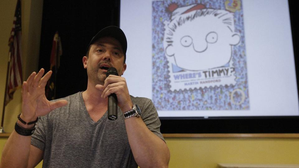 stephan pastis talking into a microphone, wearing a gray t shirt, and standing in front of a projected image of the cover of his children's book where's timmy