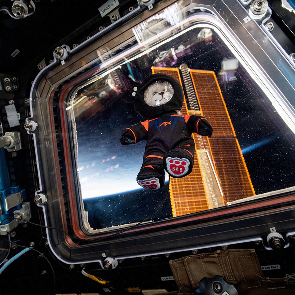 a small stuffed bear wearing a black spacesuit floats in front of a window with the distant earth in the background.