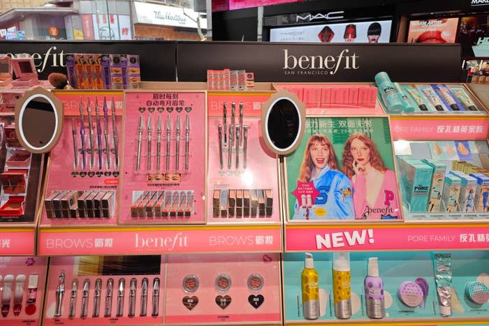 A makeup store display showcasing various Benefit products, including brow tools, blushes, and skincare items. Two women on a promotional poster