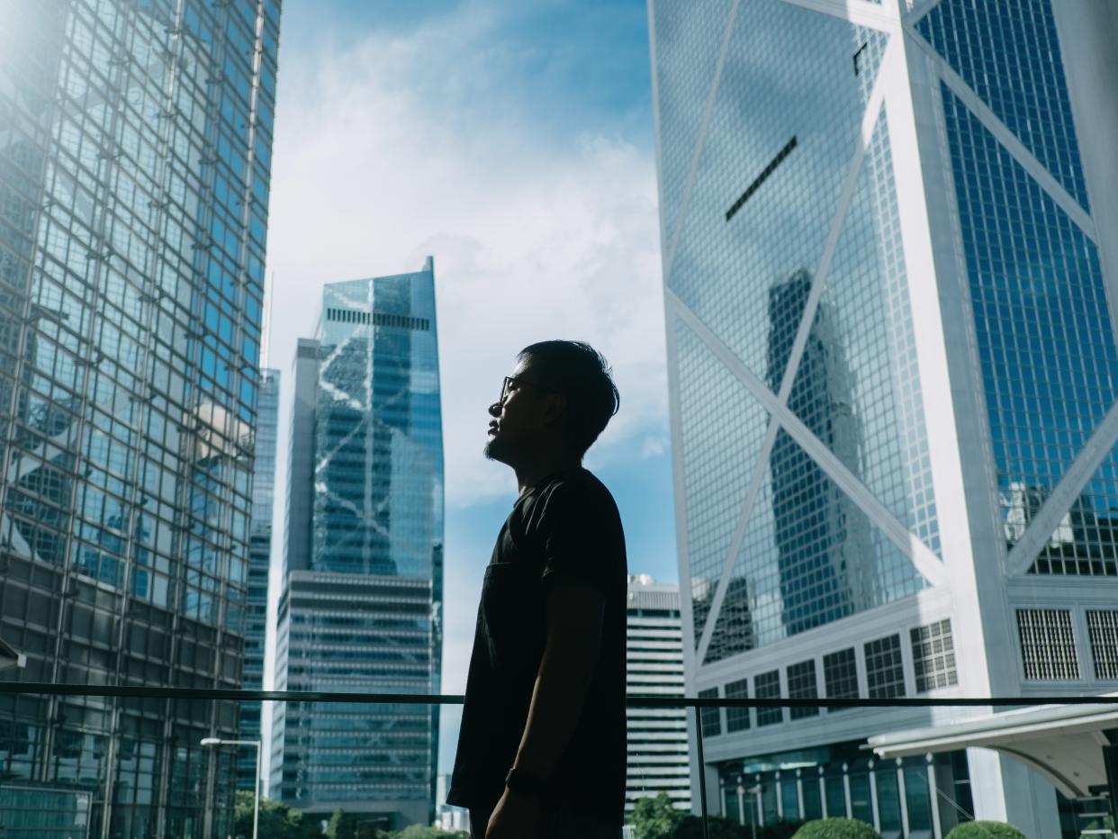 Silhouette of professional young businessman standing against contemporary financial skyscrapers and looking up into sky with positive emotion