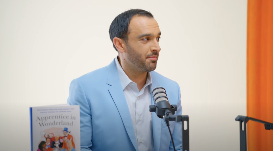 Ramin Setoodeh, in a light blazer, is sitting in front of a microphone. A book titled "Apprentice in Wonderland" is visible next to him