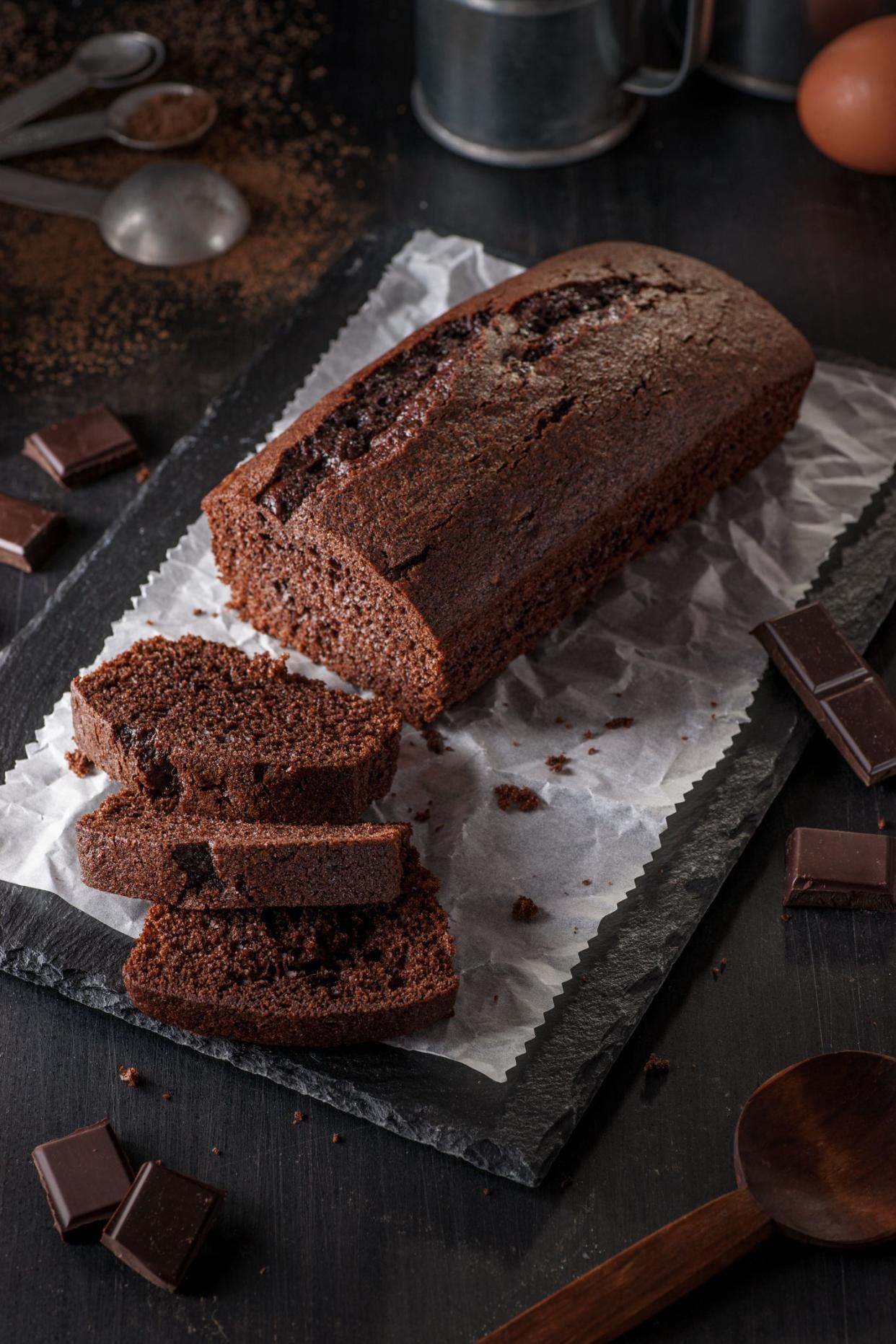 High resolution digital capture of a freshly baked chocolate loaf cake with three slices cut to reveal internal texture, sitting on a piece of wax paper on a slate slab, and surrounded by implements and ingredients used in its preparation.